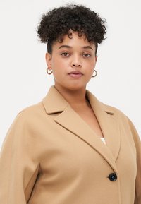 Beige oversized kabát s šálovým límcem, jedním černým knoflíkem a měkkou texturou, navržený pro uvolněný střih.