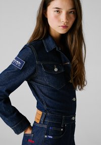 Veste en denim foncé avec un devant boutonné, des poches poitrine, des coutures claires et des patches brodés présentant un logo de course et des accents.