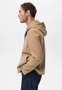 Beige hættejakke lavet af tekstureret stof, med lynlås foran og sidelommer. Designet er enkelt med en afslappet pasform og neutral farve.