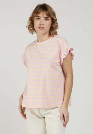 Femme aux cheveux blonds courts et ondulés, portant un t-shirt rayé rose et jaune avec des manches volantées et un pantalon de couleur crème, sur un fond uni.