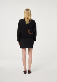 Blonde Frau mit schwarzem Pullover mit braunem Wirbelmuster, schwarzer Rock und dunkelroten Schuhen, die mit dem Rücken zur Kamera steht, vor einem einfarbigen Hintergrund.