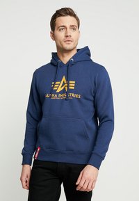 Uomo che indossa un hoodie blu di Alpha Industries con un logo arancione, in piedi contro uno sfondo chiaro e uniforme, guardando lievemente di lato.