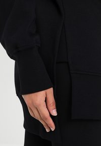 Black sweater with ribbed cuffs and hem, featuring a relaxed fit and soft texture. Hand partially visible against the garment.