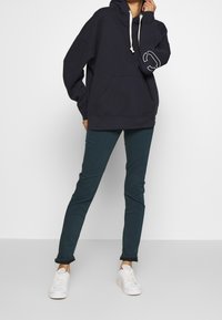 Mörkblå oversized hoodie med framficka, vita dragsnören och en logga på ärmen, kombinerad med mörkgröna slim fit-byxor och vita sneakers.