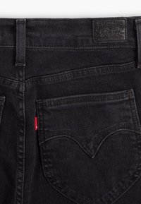 Gros plan de la poche arrière d'un jean en denim noir avec surpiqûres signature, passants pour ceinture et une petite étiquette en tissu rouge sur la couture latérale.