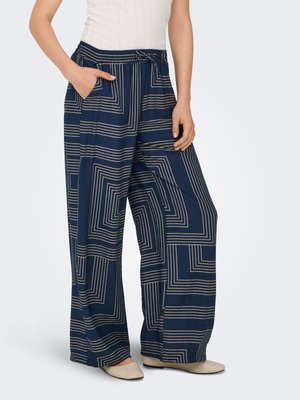Femme portant un pantalon large bleu marine avec des motifs carrés géométriques beiges, un top blanc sans manches et des chaussures plates beiges.
