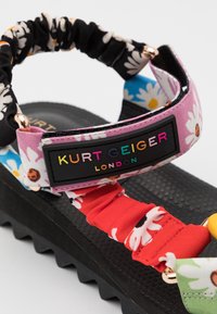 Masivní černá podešev sandálů s vícebarevnými květinovými textilními pásky a černou nášivkou s nápisem "Kurt Geiger London" v duhových písmenkách.