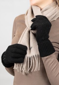Zwarte gebreide handschoenen met geribde manchetten en sprankelende accenten, gecombineerd met een lichtbeige sjaal met franjes, gedragen over een bruin bovenstuk.