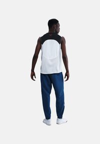 Witte mouwloze sporttop met zwarte schouders, gecombineerd met marineblauwe broek. De stof lijkt lichtgewicht en ademend, geschikt voor sport.