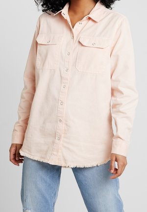 Mujer con una camisa de botones color rosa claro, con bolsillos delanteros y bajo deshilachado, combinada con jeans azules desgastados.