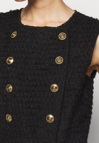 Haut sans manches texturé noir avec six boutons décoratifs en or disposés en deux rangées verticales à l'avant.