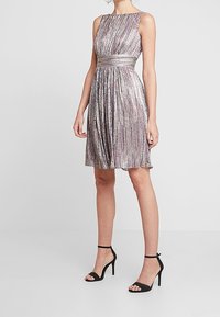 Une robe sans manches, arrivant au genou, avec une jupe évasée, arborant un motif rayé métallique dans des teintes de rose et d'argent, avec une taille cintrée.