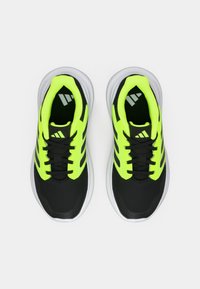 Sorte sportsko med neonlyse gule detaljer, mesh-materiale, tykke hvite såler og svarte lisser. Har tre svarte striper på sidene.