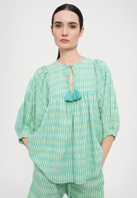 Brīvi pieguļošs, zaļš svītrainais blūze ar atslēgas caurumu kakla izgriezumā, ar tirkīza pušķiem un pūkainām piedurknēm. Izgatavota no viegla, teksturēta auduma.