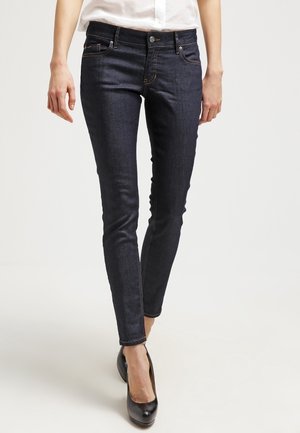 Vrouw draagt donkere skinny jeans en zwarte hoge hakken, staand tegen een effen lichte achtergrond.
