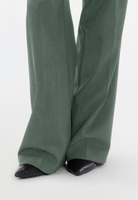 Φαρδιά παντελόνια σε απαλό πράσινο ύφασμα, με λεία υφή. Μαύρα παπούτσια με μυτερή μύτη φαίνονται στο κάτω μέρος, ολοκληρώνοντας το σύνολο.