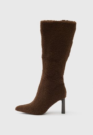 Bota con tacón marrón hecha de tela texturizada. Presenta una puntera puntiaguda y una silueta ajustada que se extiende hasta la mitad de la pantorrilla con un tacón de aguja negro.