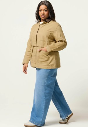 Vrouw met halflang donker haar, gekleed in een beige jas met zakken en blauwe wijd uitlopende jeans, lopend tegen een effen lichte achtergrond.