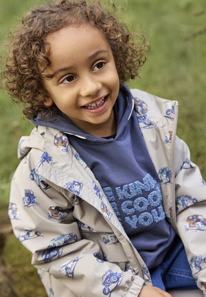 Lächendes Kind mit lockigem Haar, das einen blauen Hoodie und eine beige Jacke mit Autodrucken trägt, sitzt im Freien auf dem Gras.