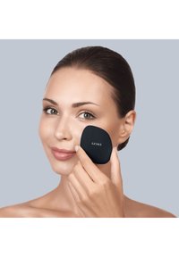 Black cosmetic applicator by GESKE held against a light complexion. Smooth texture, oval shape, with brand text visible on the surface.
