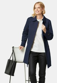 Navy blue trench coat with a button front, long sleeves, and side pockets, worn over a white top and black pants, with a black tote bag.