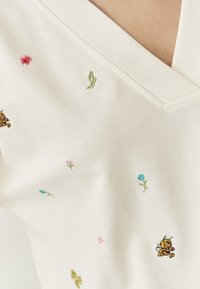 Katoenen V-hals shirt in off-wit met kleurrijke bloemen- en bijborduursels. Kenmerken zijn een zachte textuur en decoratieve stiksels langs de halslijn.
