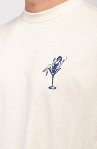 Camiseta de manga corta color blanco roto, hecha de algodón, con un gráfico azul de una figura descansando en una copa de martini en el frente.