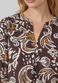 Bruine stof met een patroon van witte en gouden bloemdesigns. Heeft een v-halslijn en een zachte textuur. Geschikt voor casual kleding.