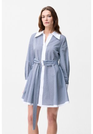 Vrouw draagt een blauw-wit verticaal gestreepte jurk met riem, lange mouwen en een witte kraag tegen een effen achtergrond.