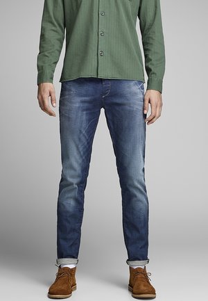 Mężczyzna ubrany w wyblakłe niebieskie obcisłe jeansy, zieloną koszulę na guziki z długimi rękawami, białe skarpetki i brązowe zamszowe buty sznurowane.