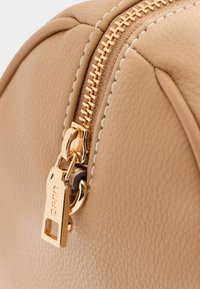 Detailní záběr béžové kožené tašky se zlatým kovovým zipem a táhlem, s kontrastním prošíváním podél okrajů. Hladký povrch a zaoblený tvar.