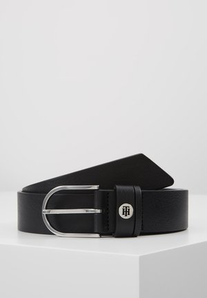 Ceinture en cuir noir avec boucle en métal argenté et petit détail de logo rond sur la boucle de ceinture, posée sur une surface blanche.