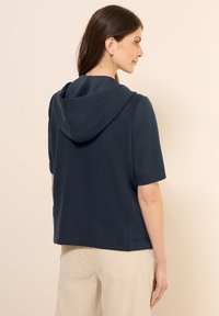 Navy Kapuzenoberteil mit kurzen Ärmeln, das einen lockeren Schnitt und eine weiche Textur bietet. Verfügt über eine Kängurutasche und eine Ziernaht am Saum.