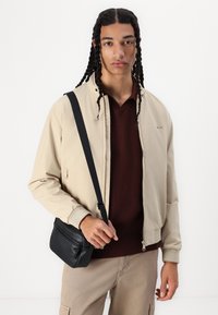Veste bomber beige avec fermeture éclair, poignets côtelés et poches. Portée par-dessus un pull en tricot bordeaux, accompagnée d'un petit sac bandoulière noir.