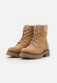 Botas de tobillo de cuero marrón con forro de piel de oveja, seis ojales metálicos y cordones beige. Presenta una suela de goma resistente y sutiles detalles de tela gris.