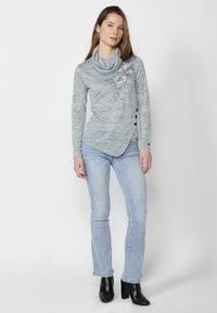 Haut en tricot gris avec un col drapé, ornements floraux sur la gauche, et accents de boutons noirs. Associé à un jean bleu clair et des bottines noires.