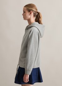 Grau zipfelige Kapuzenjacke mit einer geräumigen Kapuze und Kordelzug, die neben einem Jeansrock präsentiert wird. Der Stoff scheint weich mit einer glatten Textur zu sein.