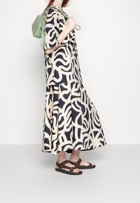 Completo in beige e blu con motivo, composto da top e gonna; tessuto leggero, vestibilità rilassata. Indossato con sandali marroni con cinturini e una borsa a tracolla verde.