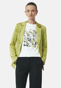 Lichtgroene satijnen blazer over een wit T-shirt met een bloemenontwerp in geel, zwart en blauw, gecombineerd met zwarte broek.