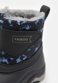 Svart vinterstövel med mönstrad tyg som har blå byggfordon. Grå fluffig foder och en sidologotyp är synliga.