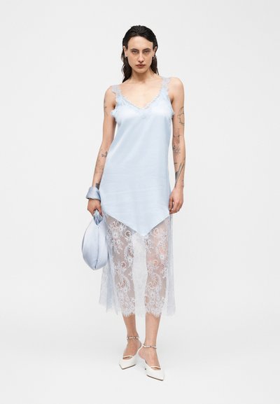 Γυναίκα με τατουάζ που φοράει ανοιχτό μπλε φόρεμα με δαντελένιο τελείωμα, κρατώντας τσάντα σετ και φοράει λευκές γόβες με μυτερές μύτες και λουράκια στον αστράγαλο.