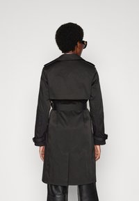 Svart trench coat med bälte i midjan, hög krage och flikdetalj bak. Tillverkad av ett mjukt, vattenavvisande tyg.