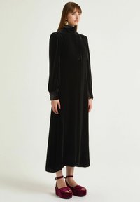 Abito in velluto nero con colletto alto, maniche lunghe e dettaglio con bottoni. Abbinato a scarpe platform bordeaux con cinturino alla caviglia.