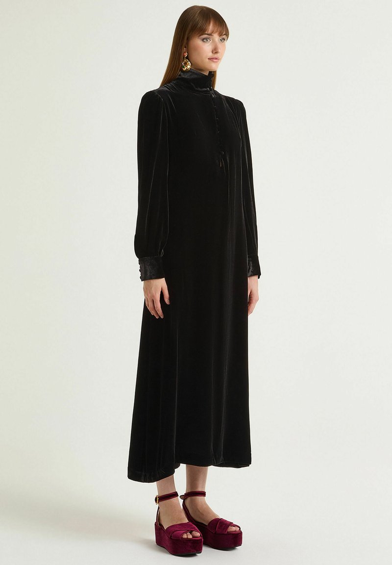 Abito in velluto nero con colletto alto, maniche lunghe e dettaglio con bottoni. Abbinato a scarpe platform bordeaux con cinturino alla caviglia.