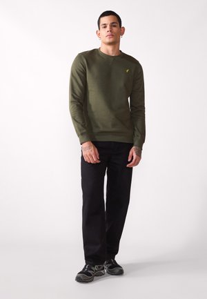 Maglione verde oliva con colletto rotondo e polsini a costine, indossato con jeans neri e scarpe da ginnastica, con un piccolo logo giallo sul petto.