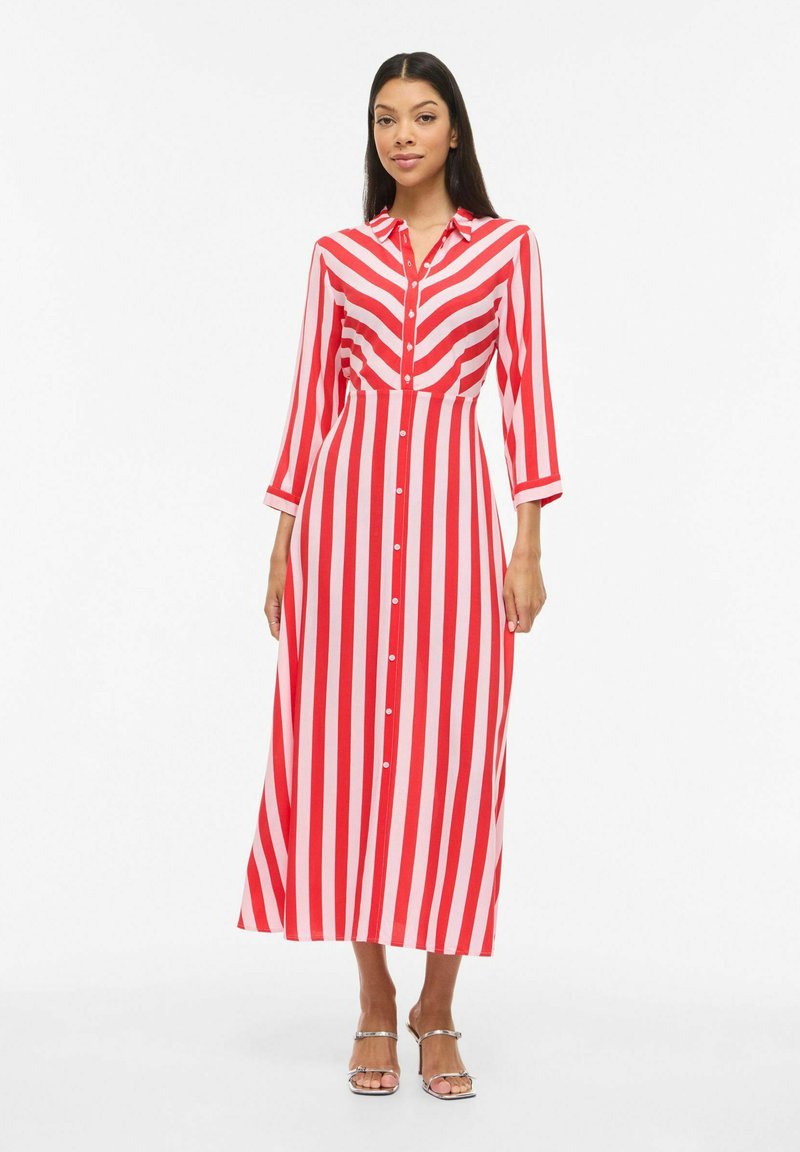 Femme portant une robe longue à boutons rayée rouge et rose à manches longues, avec des talons argentés ouverts, debout devant un fond blanc.