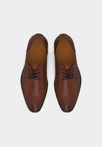 Ζευγάρι καλοχτενισμένων καφέ δερμάτινων επίσημων παπουτσιών με μαύρα κορδόνια και μαύρες σόλες, σε θέα από πάνω σε λευκό φόντο.