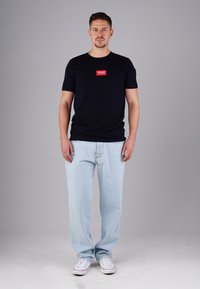 Mann, der nach vorne blickt, trägt ein schwarzes T-Shirt mit rotem Logo, hellblaue Jeans und weiße Sneaker vor einem schlichten grauen Hintergrund.