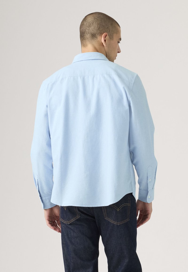 Mann trägt ein hellblaues langärmeliges Hemd und dunkelblaue Jeans, von hinten vor einem einfarbigen Hintergrund gezeigt.