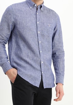 Mann trägt ein hellblaues, langärmeliges Hemd mit Brusttasche, kombiniert mit schwarzen Hosen, mit einer Hand in der Tasche.